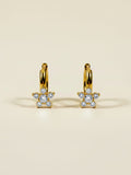 A pair of gold huggie hoop earrings with small flower-shaped clusters of clear pavé gemstones, displayed on a warm beige background.