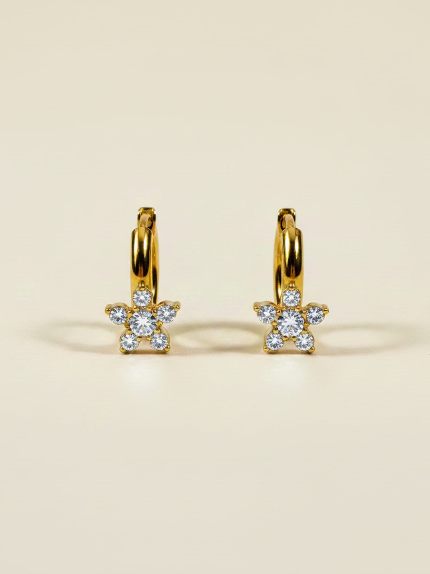 A pair of gold huggie hoop earrings with small flower-shaped clusters of clear pavé gemstones, displayed on a warm beige background.