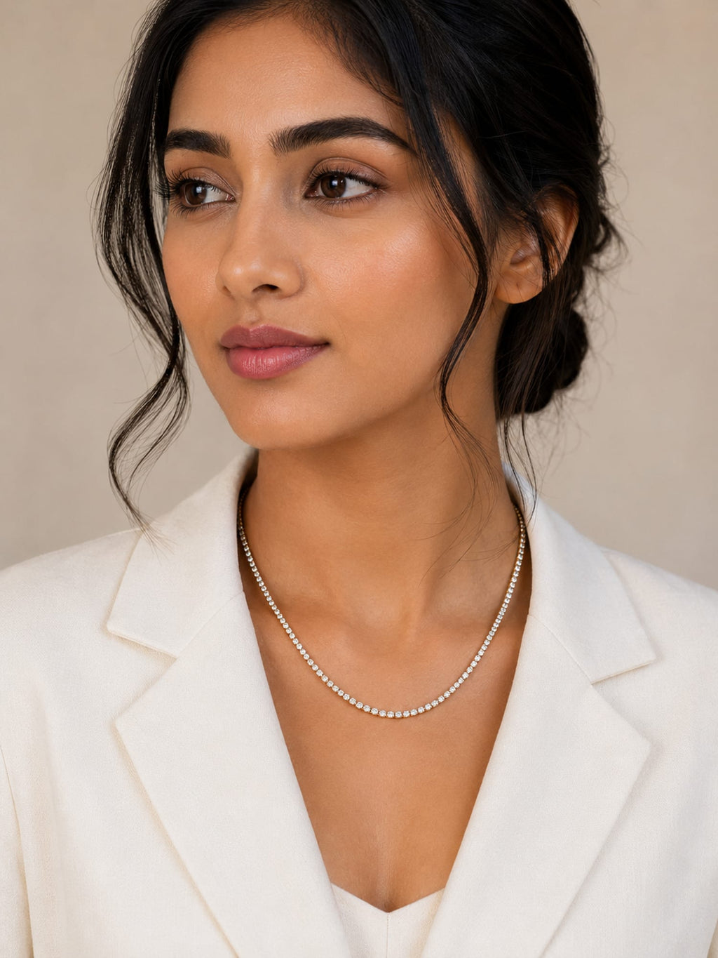 A woman with dark hair pulled back wears a delicate silver chain necklace and a white blazer against a soft beige background.