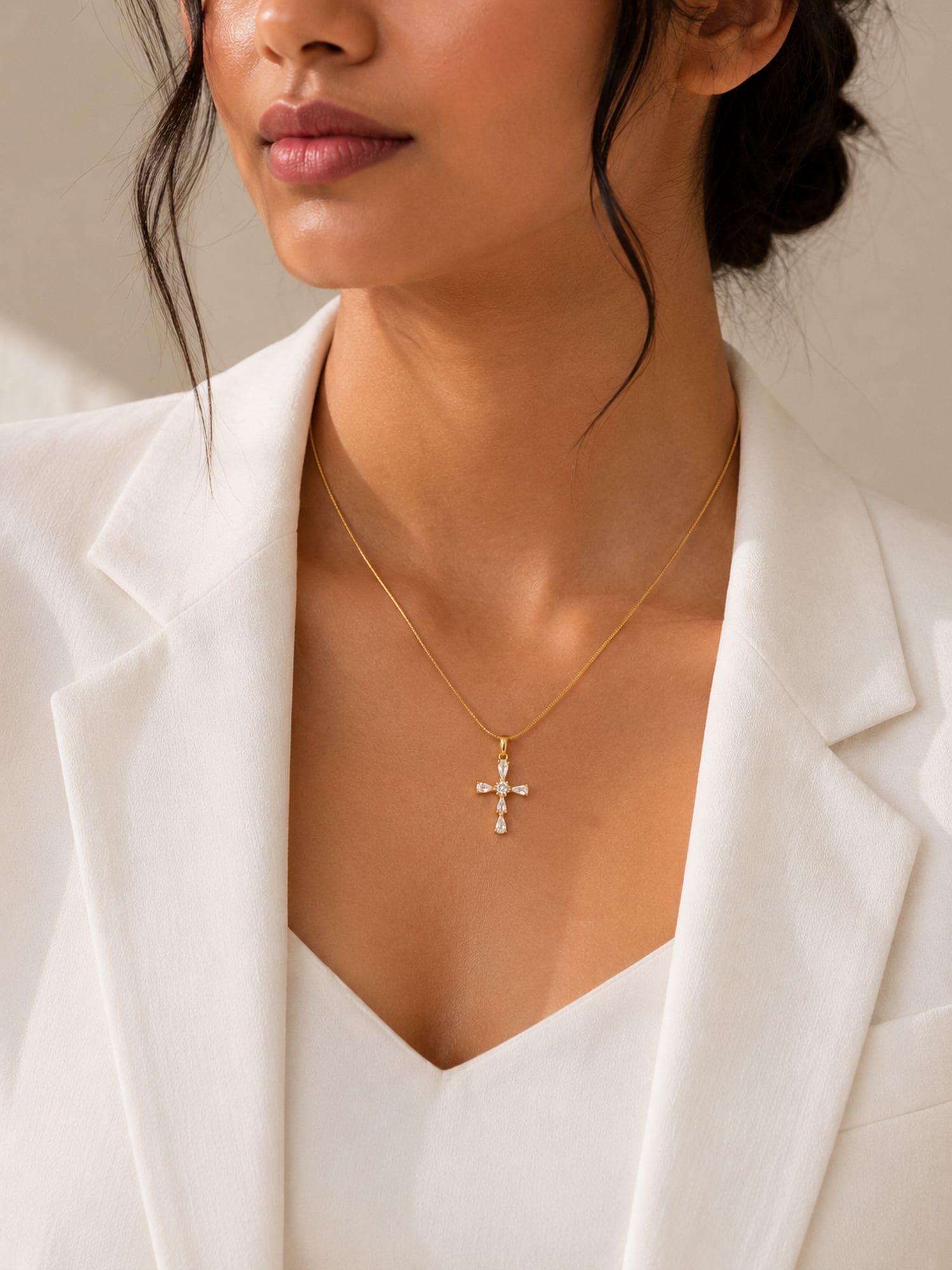 A close-up of a model wearing a delicate gold cross pendant necklace with small clear stones, styled with a white blazer.