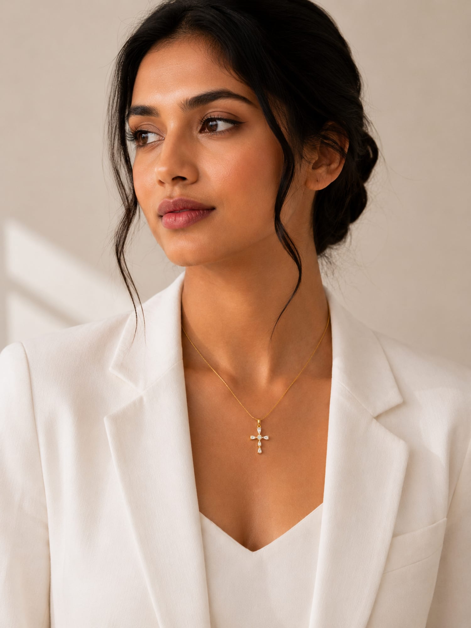 A woman with dark hair in a bun wearing a white blazer and a delicate gold cross pendant necklace, shown in a close-up portrait against a soft beige background.