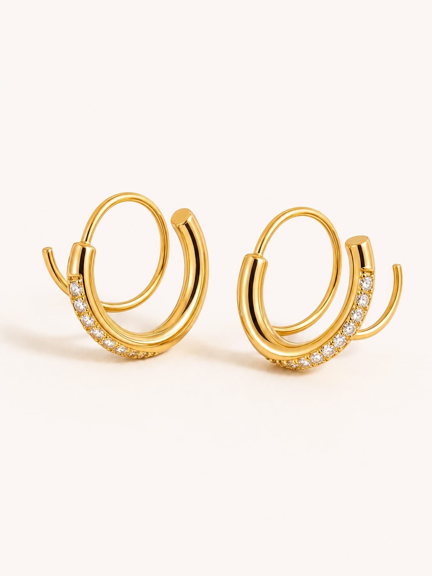 A pair of gold-toned double-hoop earrings with a row of small clear stones along the front curve, shown on a plain light beige background.