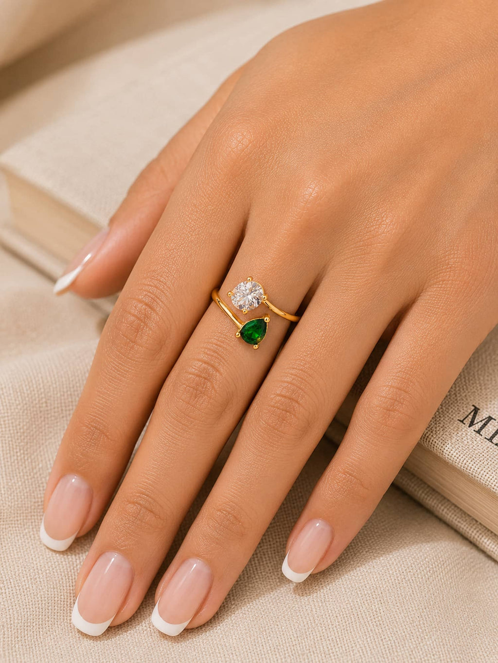 A close-up of a hand with a French manicure wearing a gold ring with a round clear gemstone and a pear-cut green gemstone, resting on a beige fabric surface next to an open book.