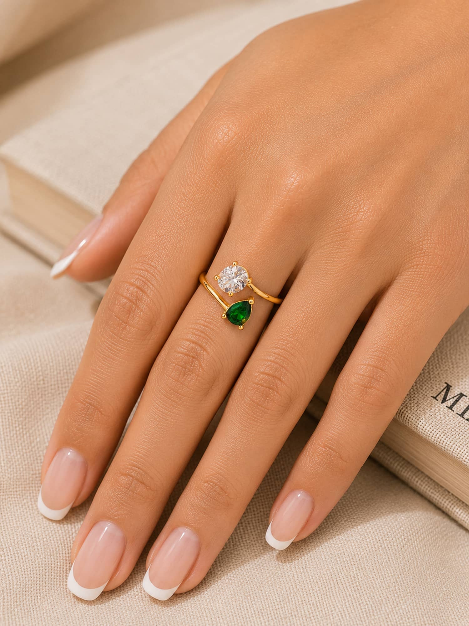 A close-up of a hand with a French manicure wearing a gold ring with a round clear gemstone and a pear-cut green gemstone, resting on a beige fabric surface next to an open book.