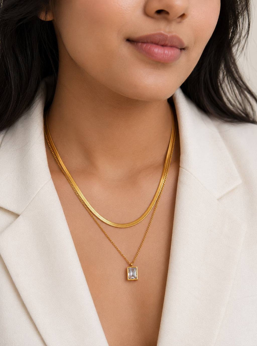 A close-up of a model wearing a double-layered gold necklace, featuring a thicker snake chain and a delicate chain with a rectangular clear gemstone pendant, styled with a white blazer.