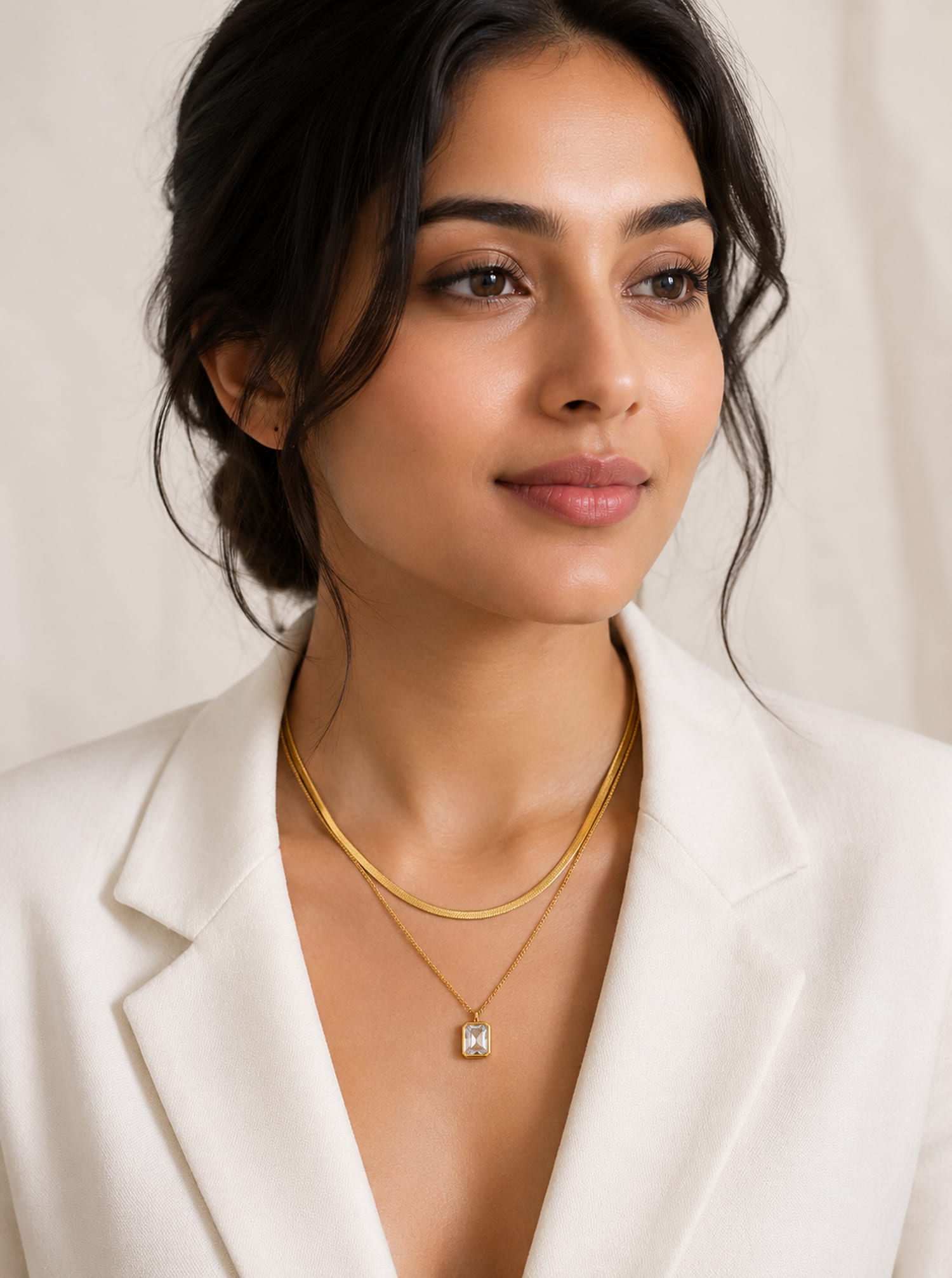 A woman wearing a double-layered gold necklace with a small square emerald cut cubic zirconia pendant, styled with a white blazer against a soft beige background.