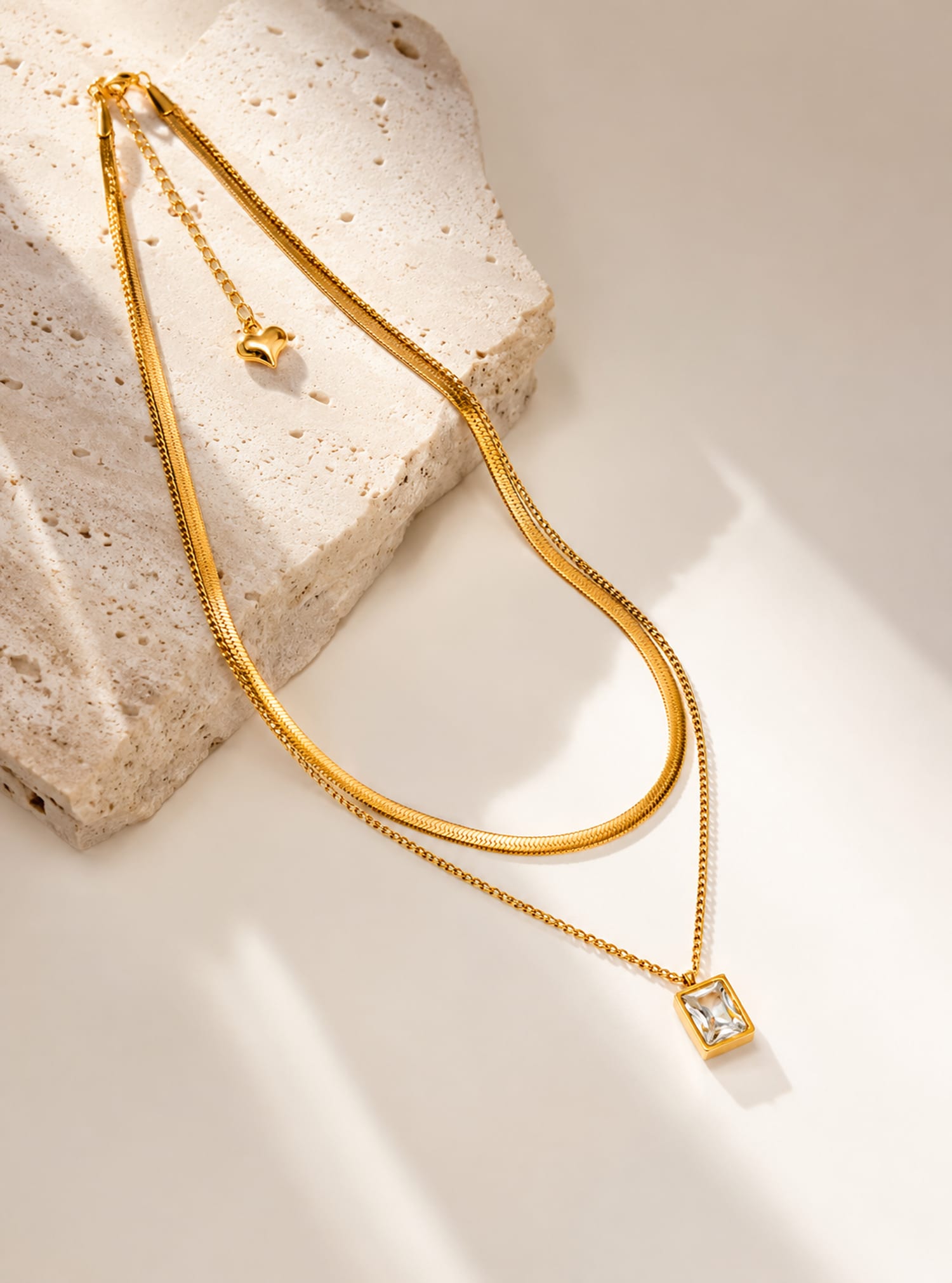 Minimalist layered snake chain necklace with emerald cut CZ pendant flat lay on textured stone surface