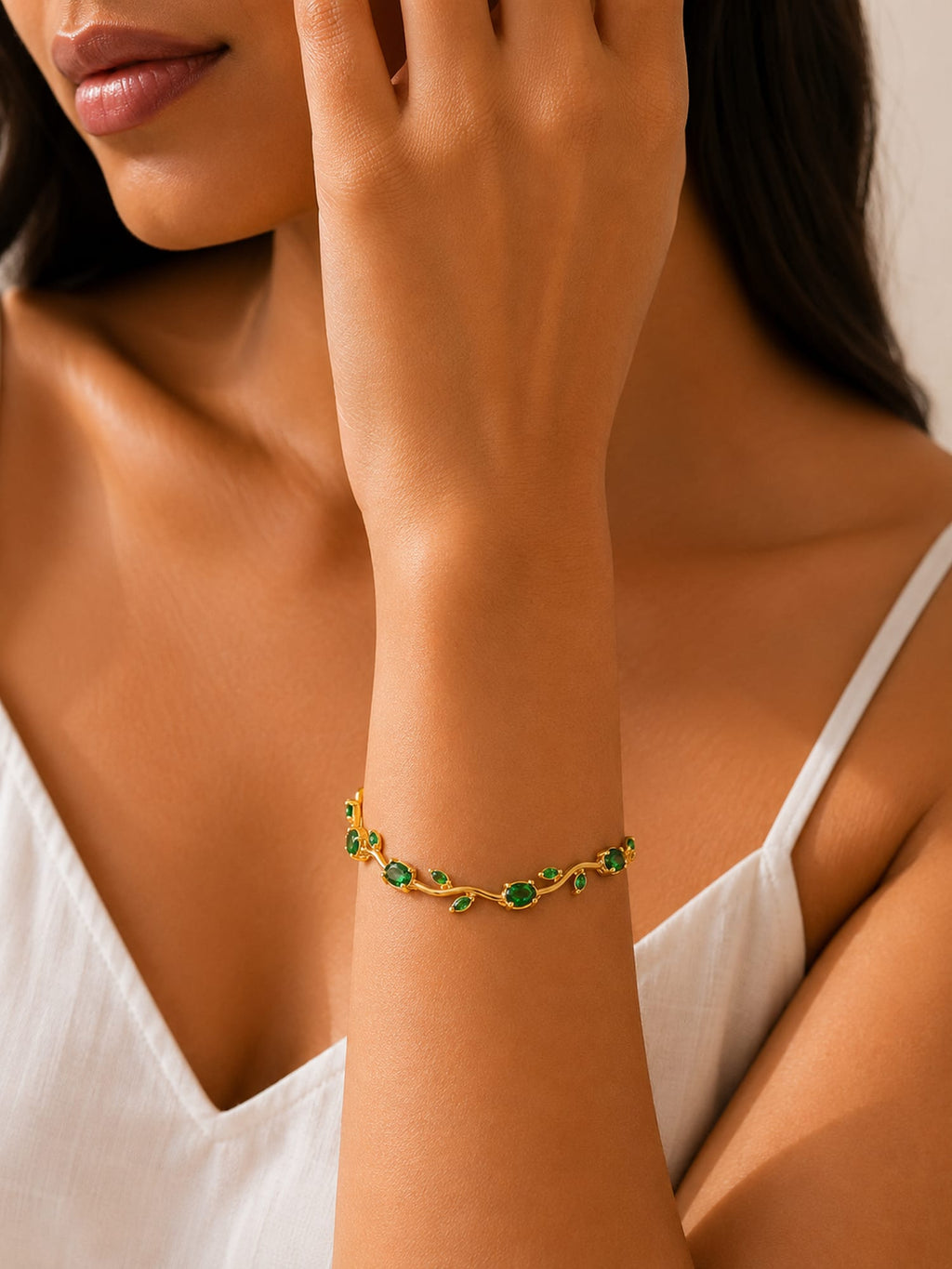 A close-up of a woman wearing a delicate gold bracelet adorned with small green gemstones on her wrist, paired with a white spaghetti-strap top.