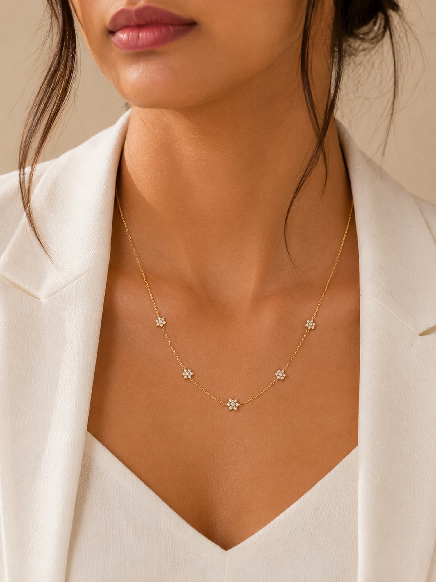 A close-up of a model wearing a delicate gold necklace with five small flower-shaped charms, styled with a white blazer.
