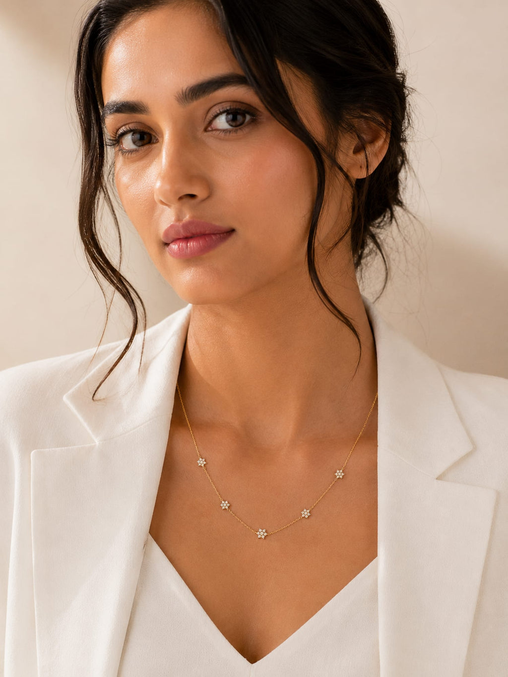A woman wearing a delicate gold necklace with small star-shaped charms, styled with a white blazer against a warm beige background.