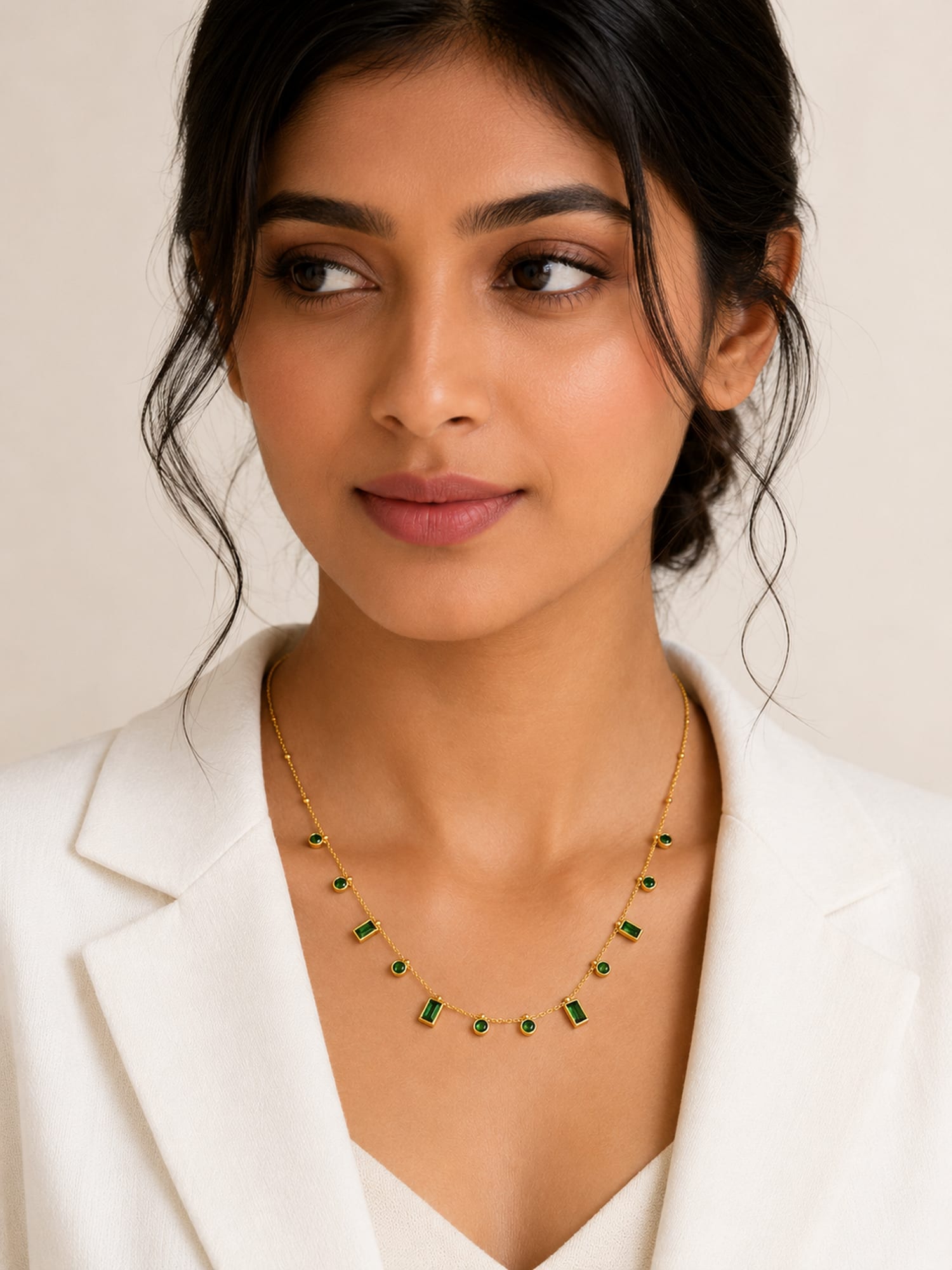 A woman wearing a delicate gold chain necklace with small green rectangular stones, styled with a white blazer against a light beige background.