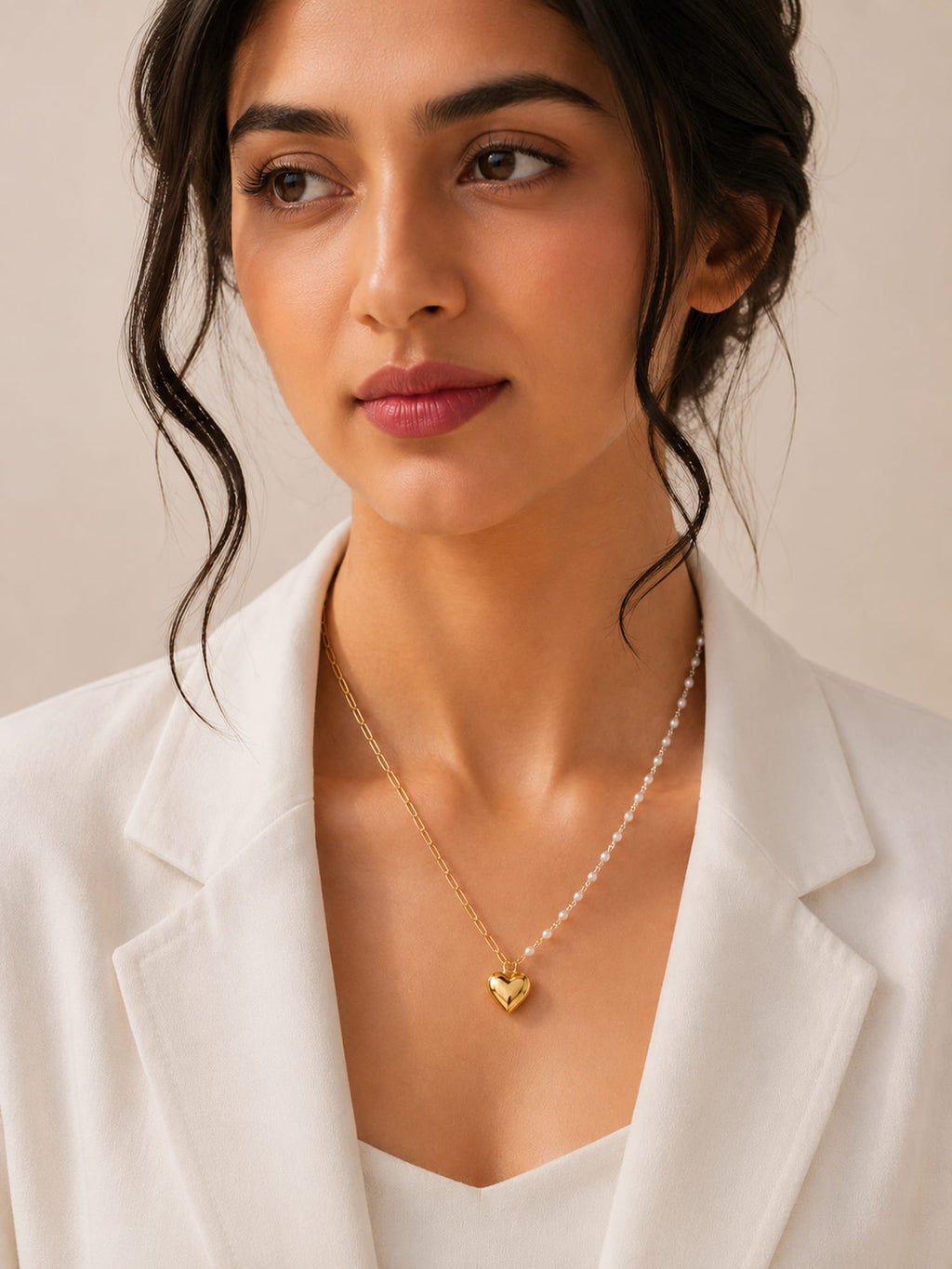 A woman wearing a delicate gold heart pendant necklace with a pearl chain, styled with a white blazer against a soft beige background.