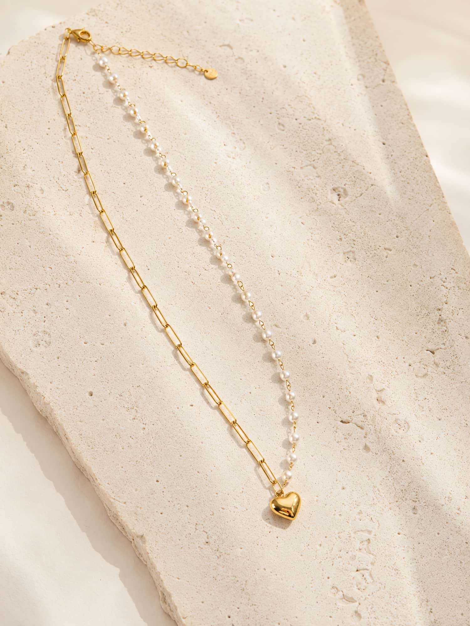 Minimalist half pearl heart pendant necklace displayed on a textured stone surface with an asymmetrical paperclip chain.