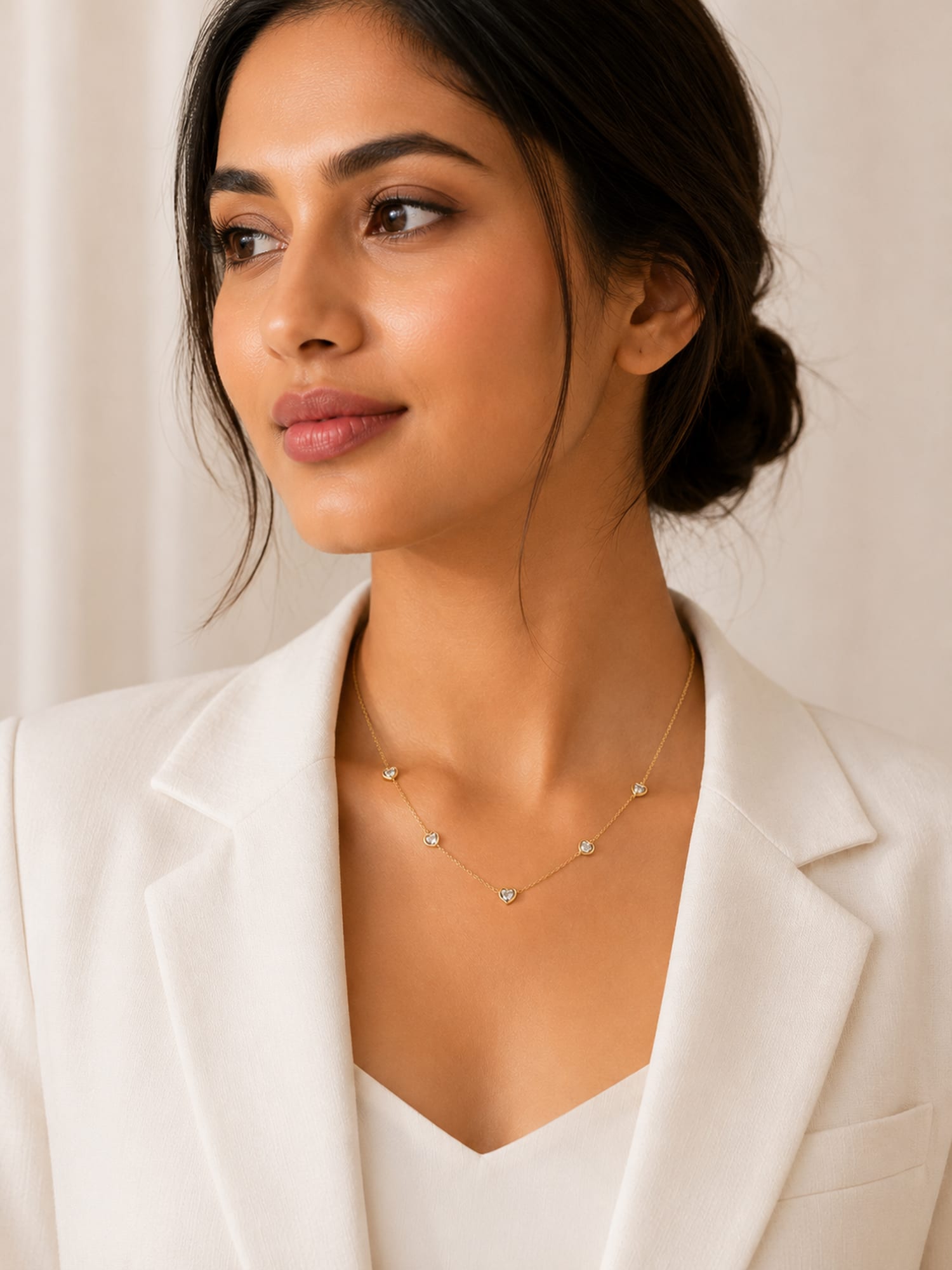 A woman wearing a delicate gold necklace with small, spaced round beads, styled with a white blazer against a soft beige background.