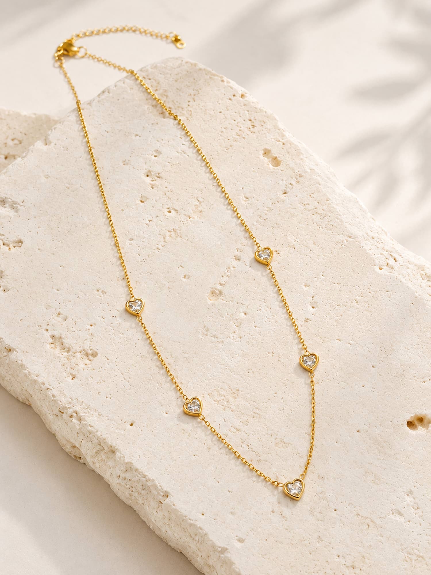 Heart station necklace with five sparkling CZ stones displayed on a textured stone surface in a gold finish.
