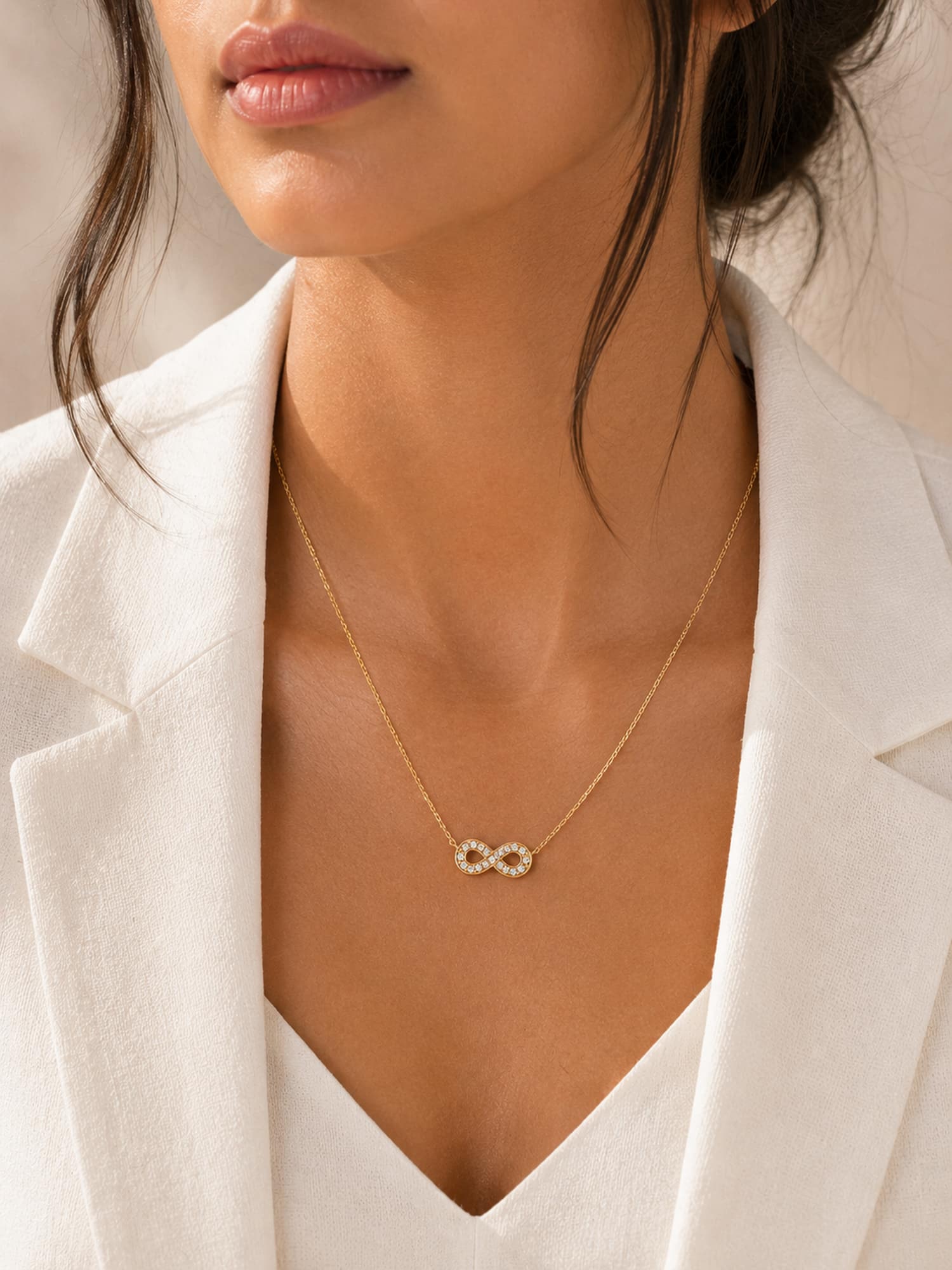 A close-up of a model wearing a delicate gold infinity necklace with a small pavé center, styled with a white blazer.
