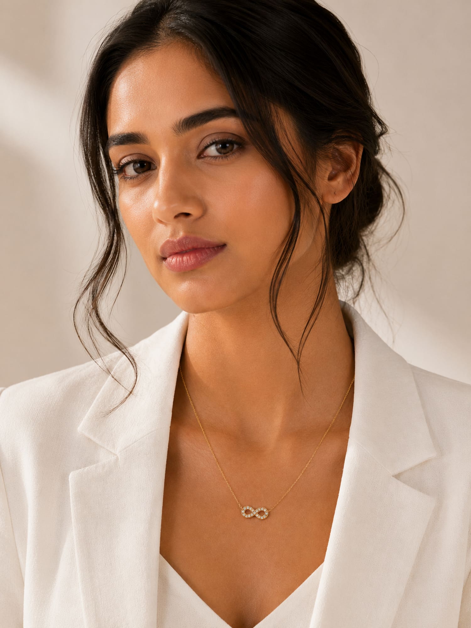 A woman wearing a delicate gold infinity necklace with a small infinity symbol pendant, styled with a white blazer against a soft beige background.