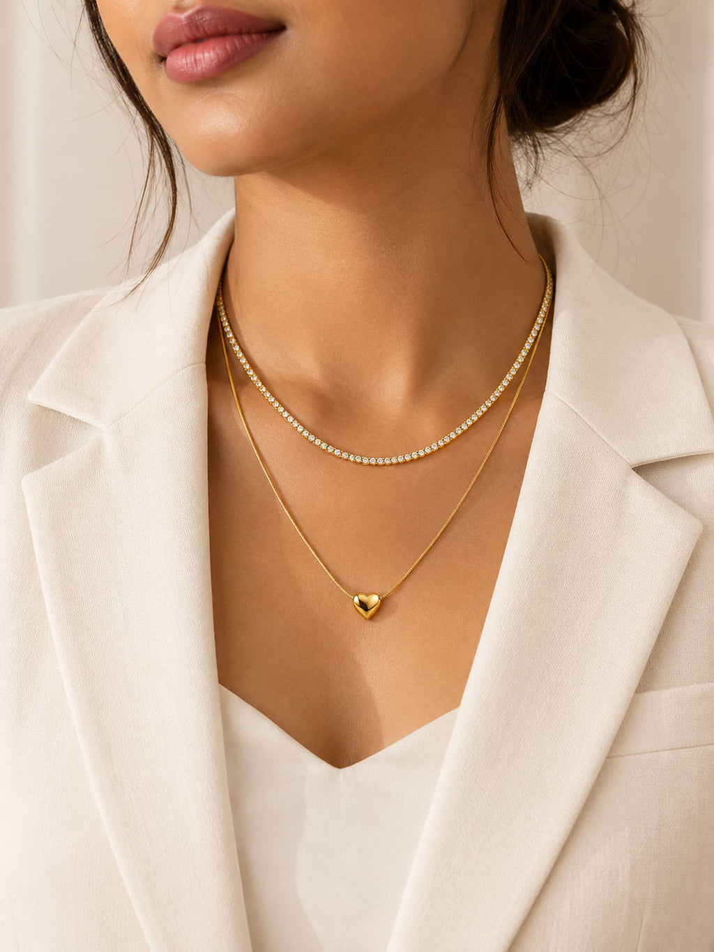A close-up of a model wearing a double-layered gold necklace with a delicate chain and a small heart pendant, styled with a white blazer.