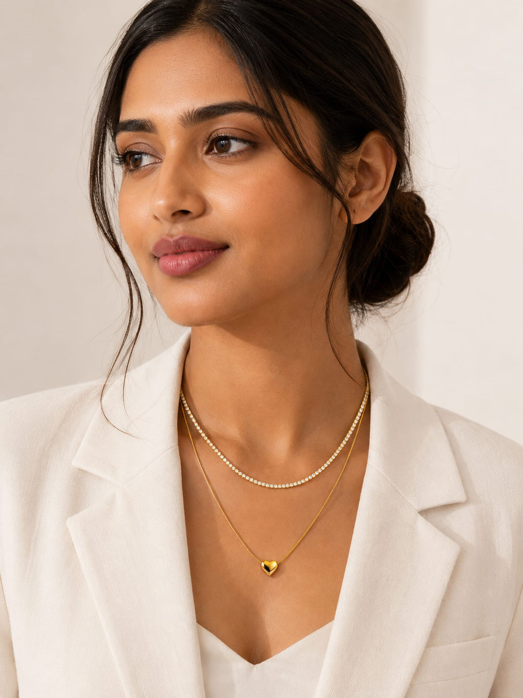 A woman wearing a double-layered necklace set, featuring a delicate gold chain with a small heart pendant and a thin pearl strand, paired with a white blazer.