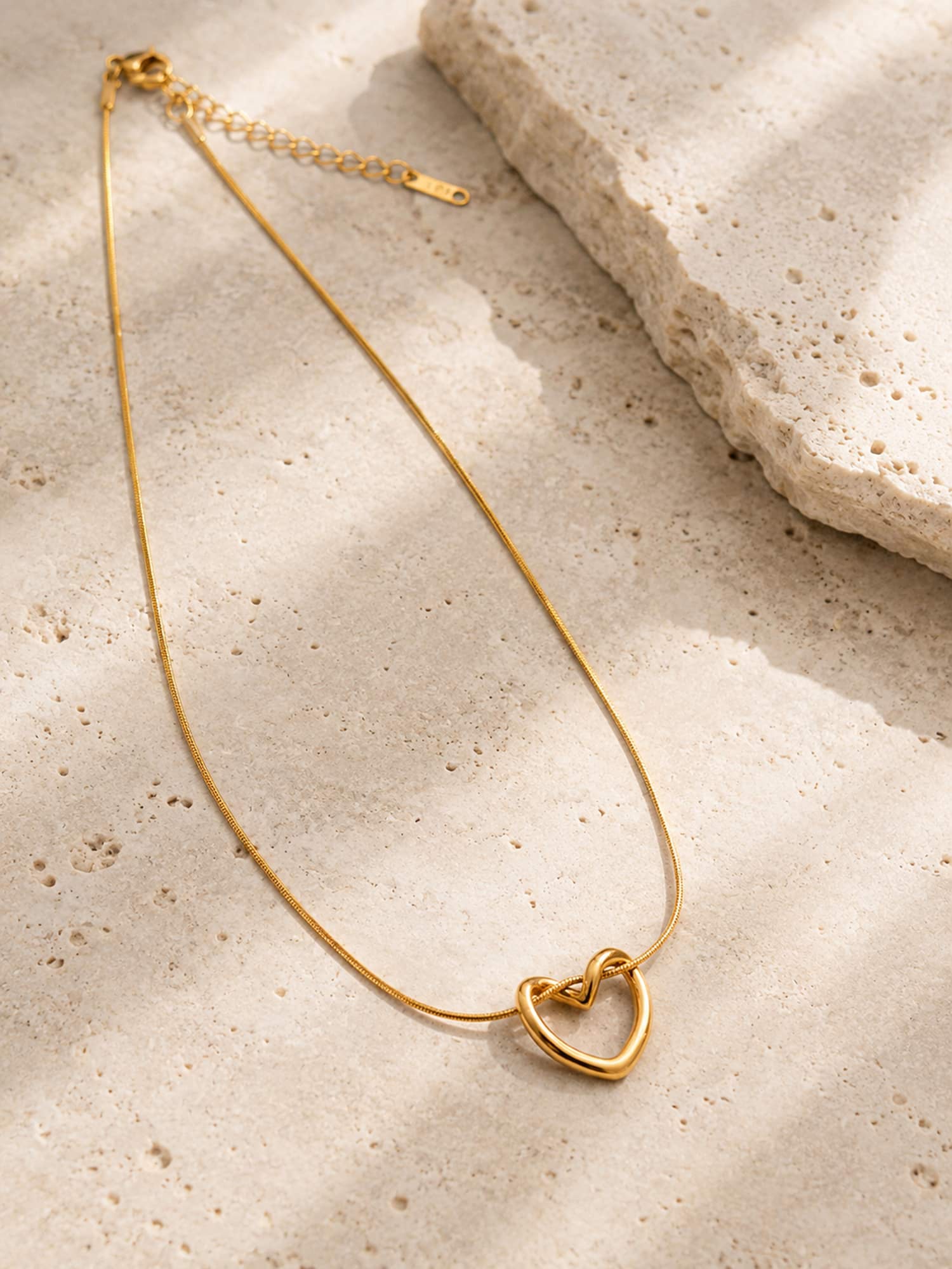 A minimalist gold chain necklace with a open-heart pendant, styled on a textured beige stone surface with soft shadows.