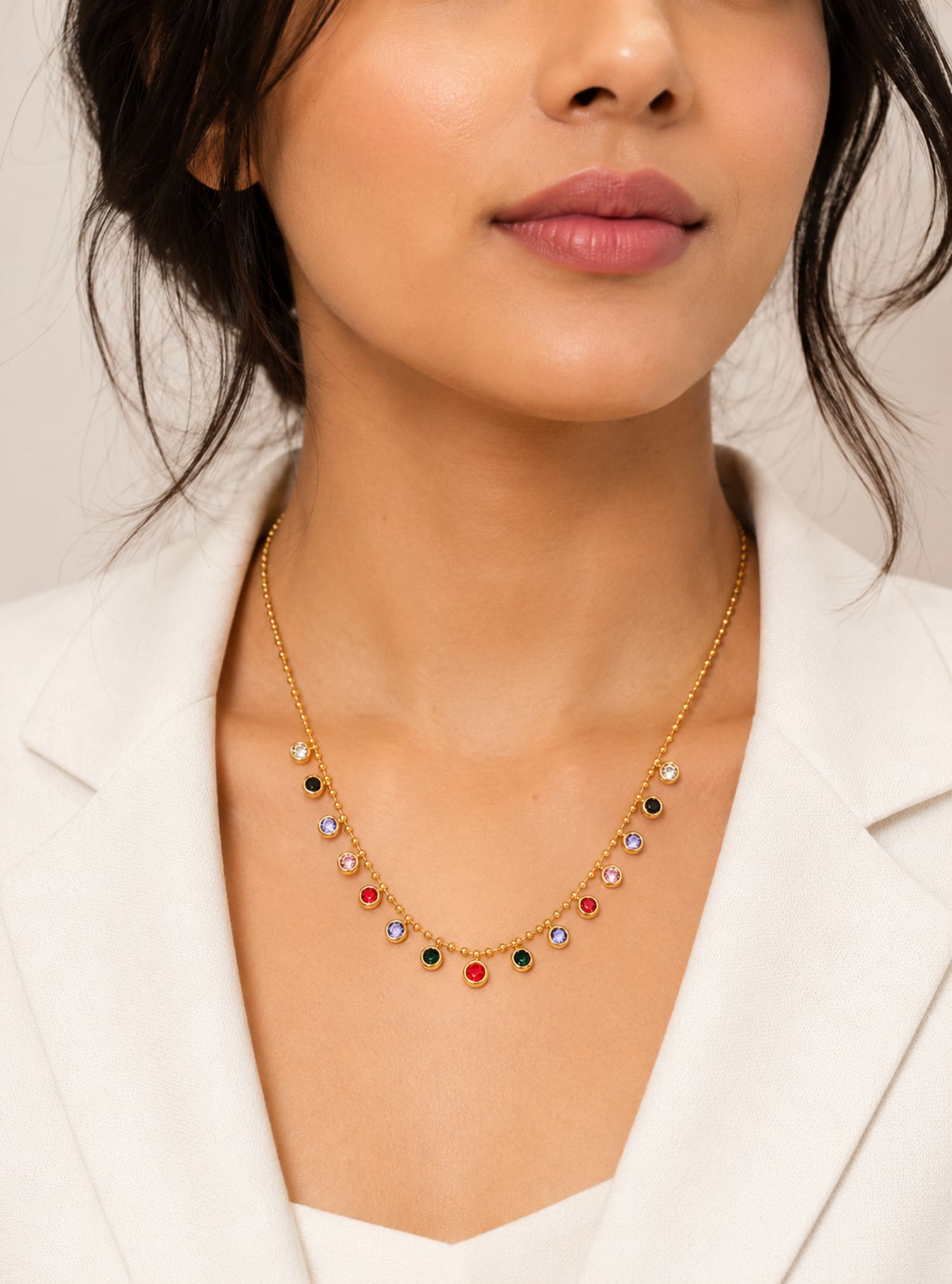 A close-up of a woman wearing a delicate gold necklace with small multicolored gemstone accents, styled with a white blazer.