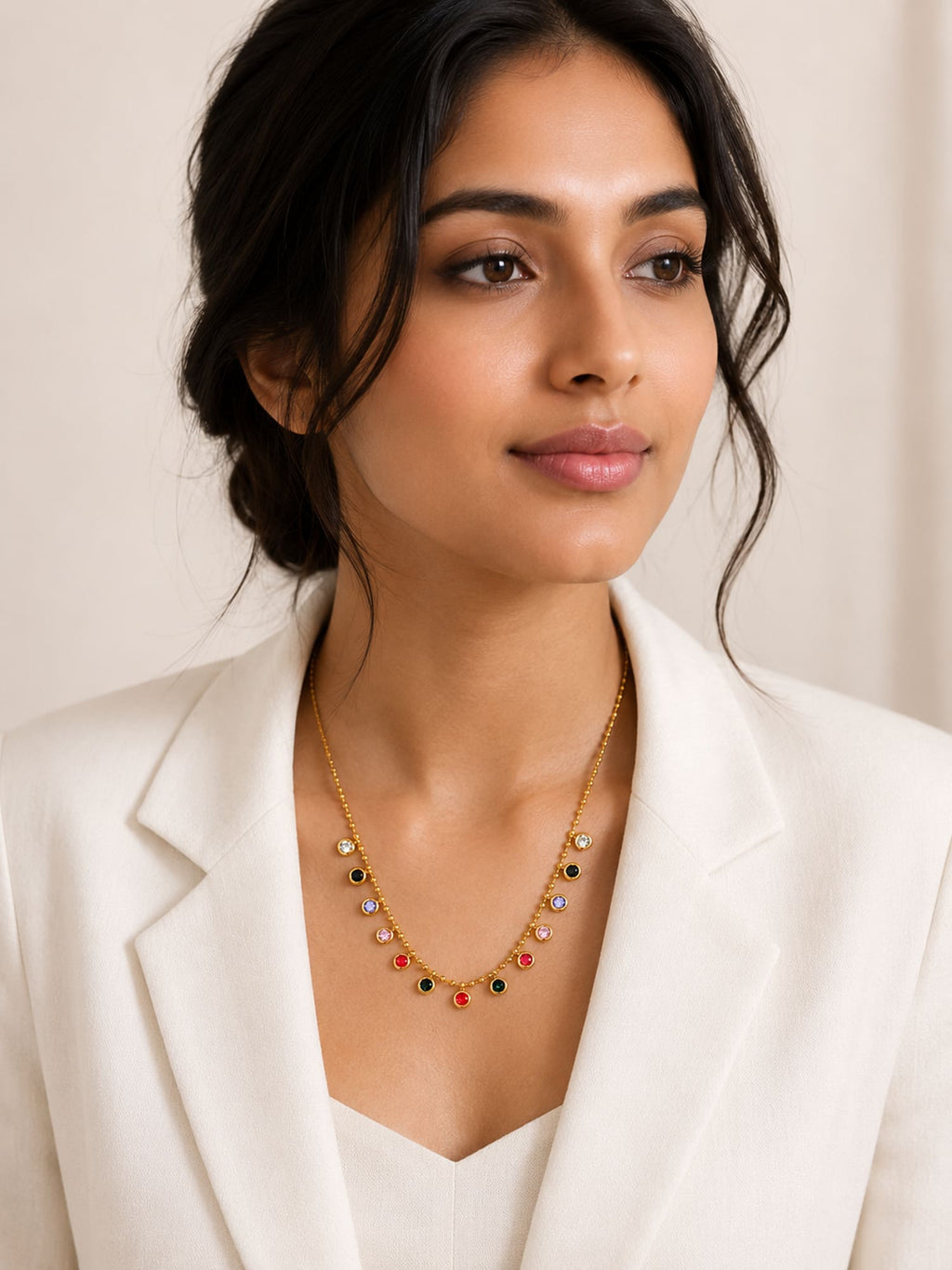A woman wearing a delicate gold necklace with small multicolored gemstone drops, styled with a white blazer against a soft beige background.