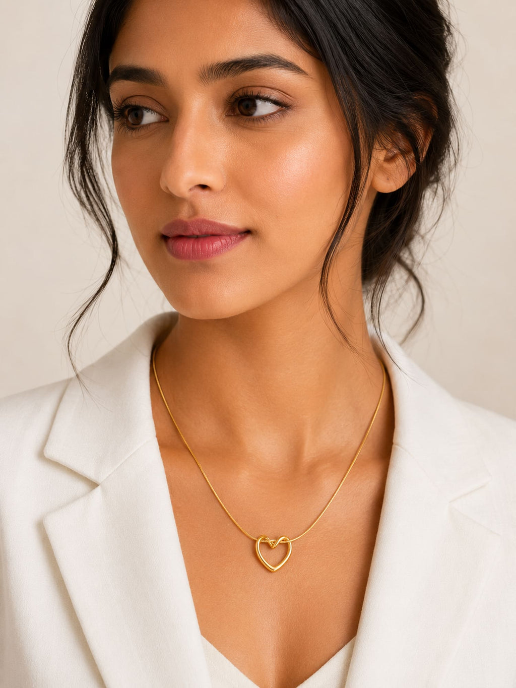 A woman wearing a delicate gold heart-shaped pendant necklace, styled with a white blazer against a soft beige background.
