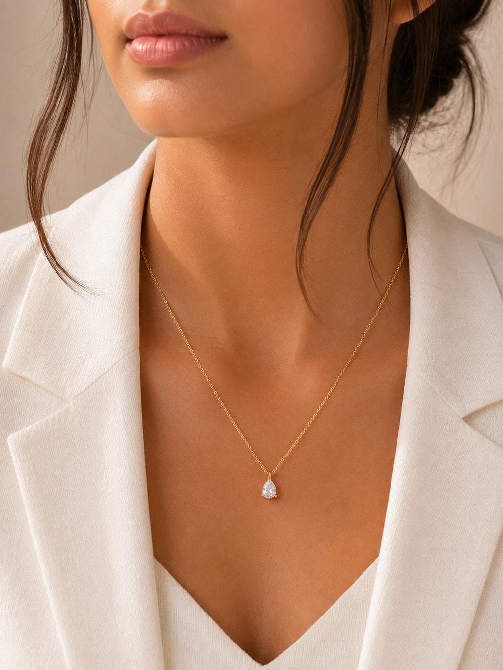 A close-up of a model wearing a delicate gold chain necklace with a small pear-cut clear gemstone pendant, styled with a white blazer.