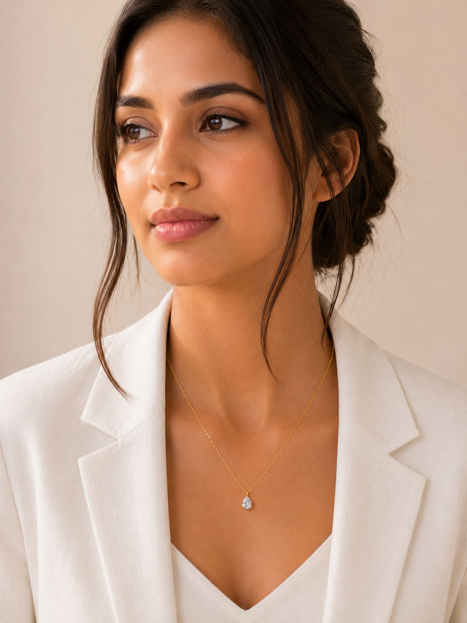 A woman wearing a delicate gold necklace with a small teardrop-shaped clear gemstone pendant, styled with a white blazer against a soft beige background.