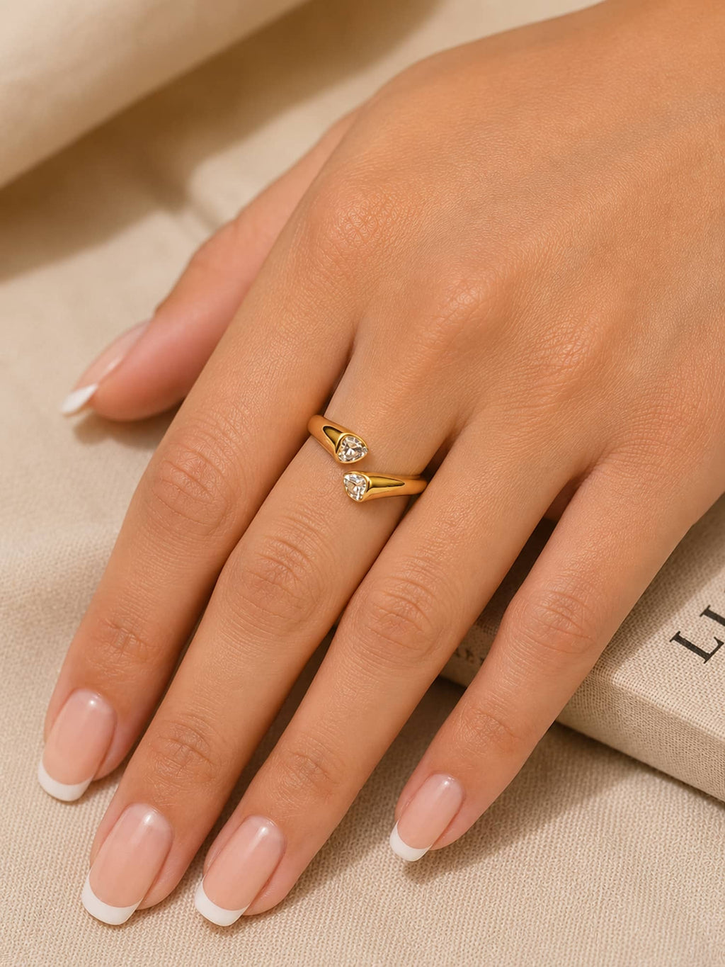 A close-up of a hand with a French manicure wearing a delicate gold ring with two small clear gemstones on a beige fabric background.