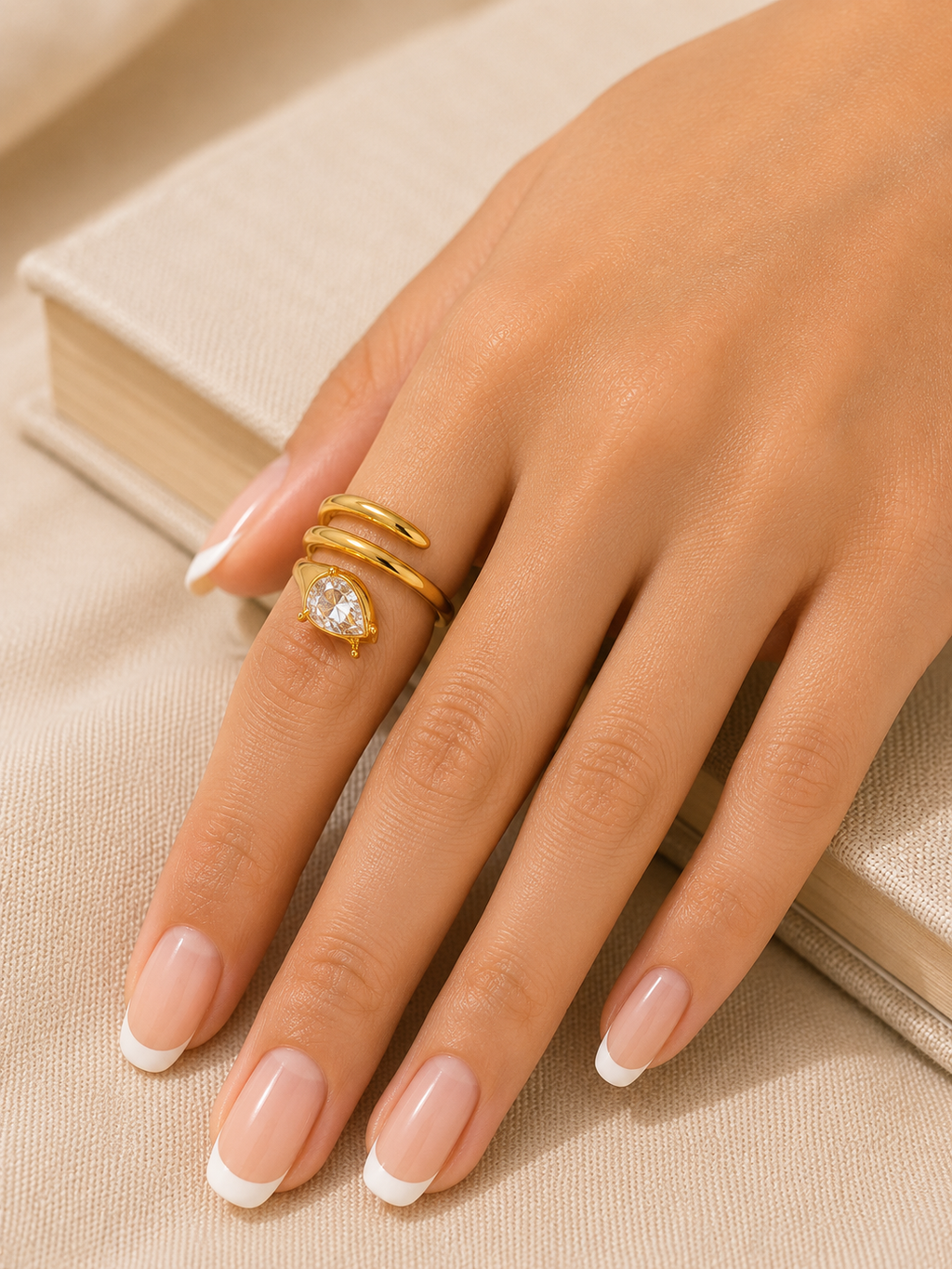 A close-up of a hand with a French manicure wearing a gold wrap ring with a heart-shaped clear gemstone, resting on a beige textured surface next to a book.